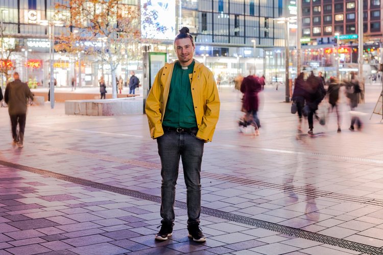 Man i grön diakonskjorta och gul jacka står på ett torg där det rör sig människor i bakgrunden.